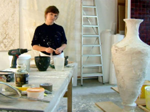 A woman stares at the camera while working in her studio. A large ceramic vessel sits in the foreground.