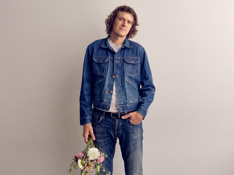 a Man with shoulder length hair in a jean jacket stands in front of a white background holding a bouquet of flowers down by his thigh