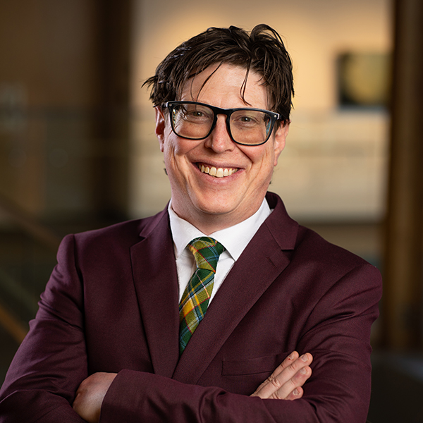 Headshot of Executive Director Steven Foord in a burgundy suit with his arms crossed and smiling. The background of paintings installed at Casa is blurred out.