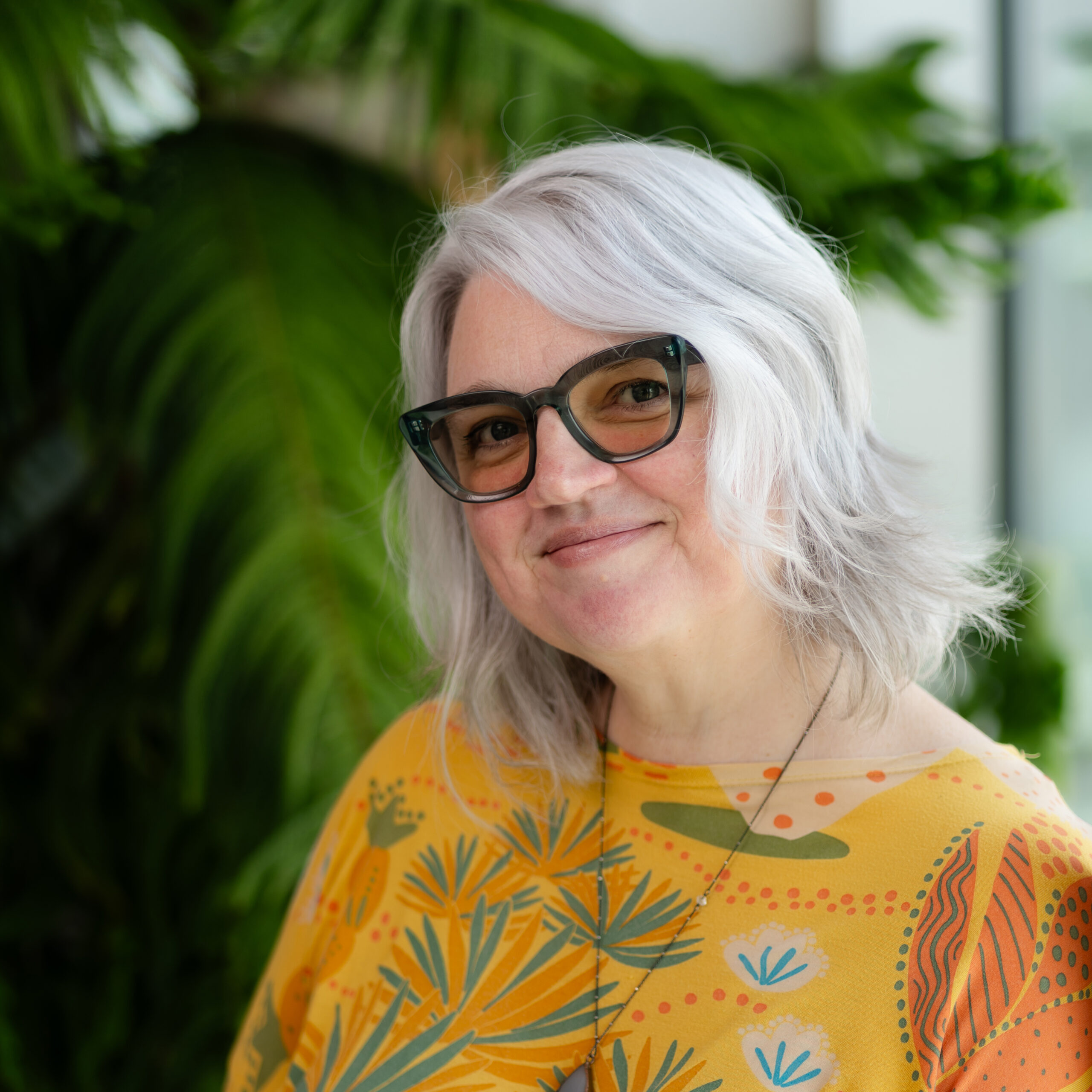 Alice Clark headshot with an amazing yellow shirt in front of an indoor pine tree at Casa.